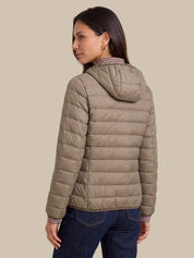 A woman shown from the back wearing a light taupe quilted puffer jacket with a hood, paired with dark blue jeans, against a solid beige background.