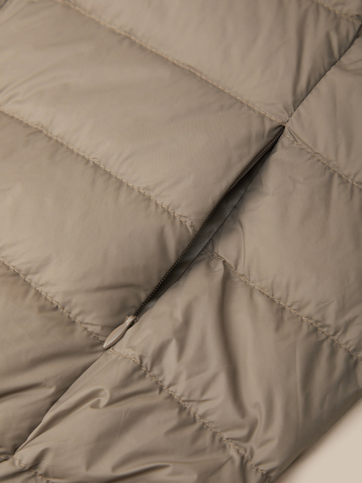 Close-up of a quilted puffer jacket in a warm taupe-beige color, showing the texture of the fabric and a black zipper running vertically.