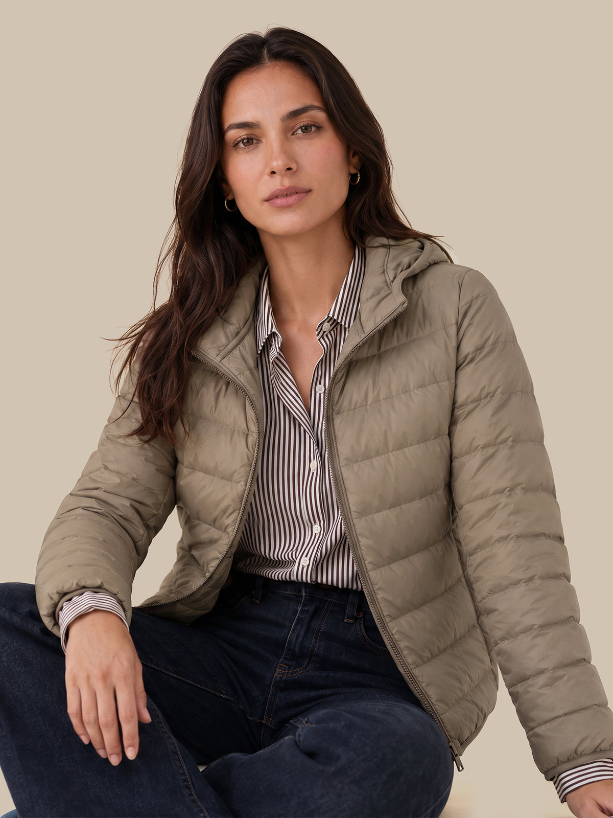 A woman wearing a light taupe quilted puffer jacket over a vertically striped button-down shirt and dark blue jeans, posed against a solid beige background.
