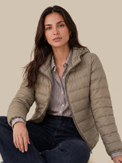 A woman wearing a light taupe quilted puffer jacket over a vertically striped button-down shirt and dark blue jeans, posed against a solid beige background.