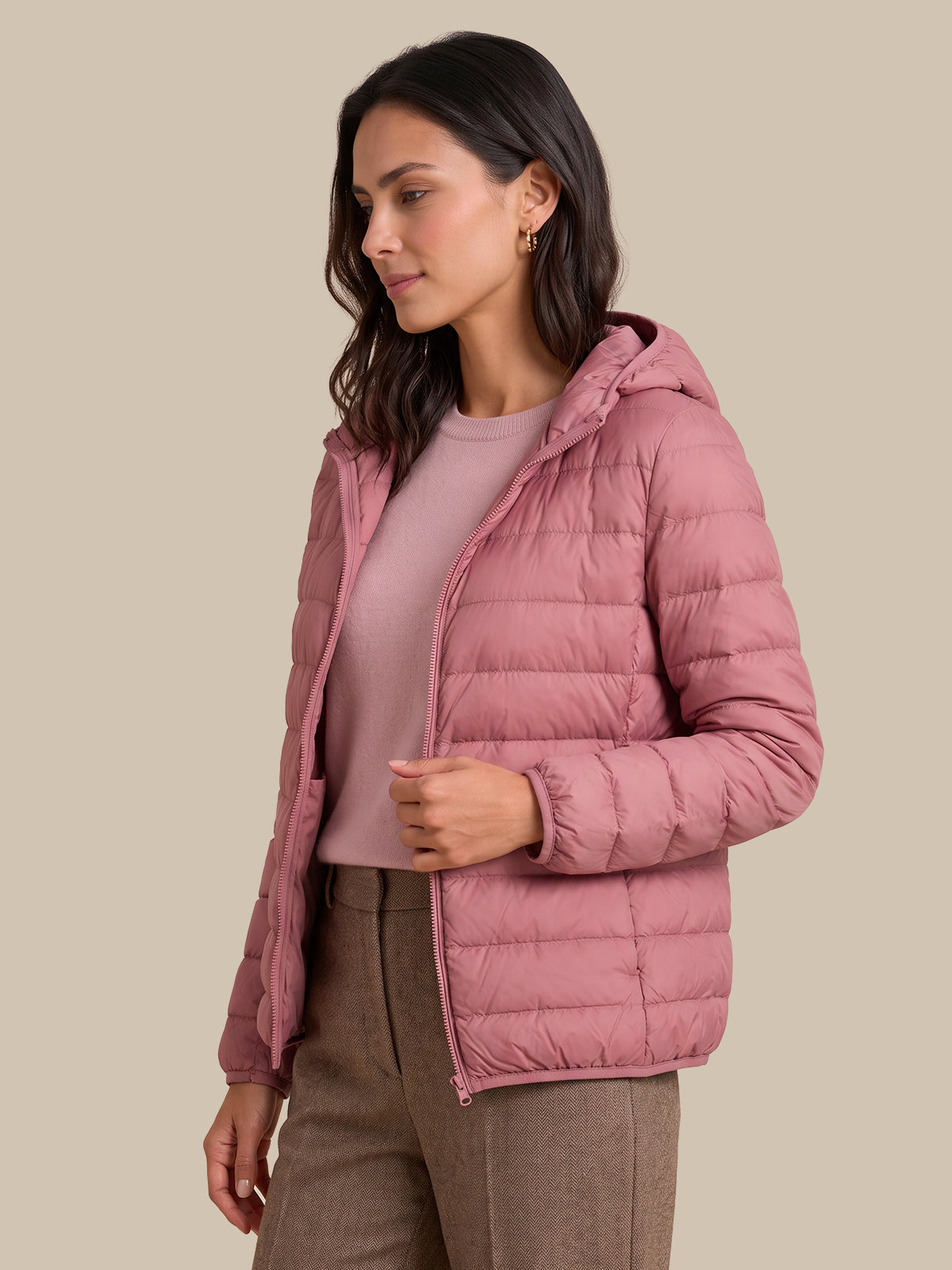 A woman wearing a dusty pink hooded puffer jacket over a matching pink top and brown trousers, photographed against a solid beige studio background.