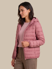 A woman wearing a dusty pink hooded puffer jacket over a matching pink top and brown trousers, photographed against a solid beige studio background.