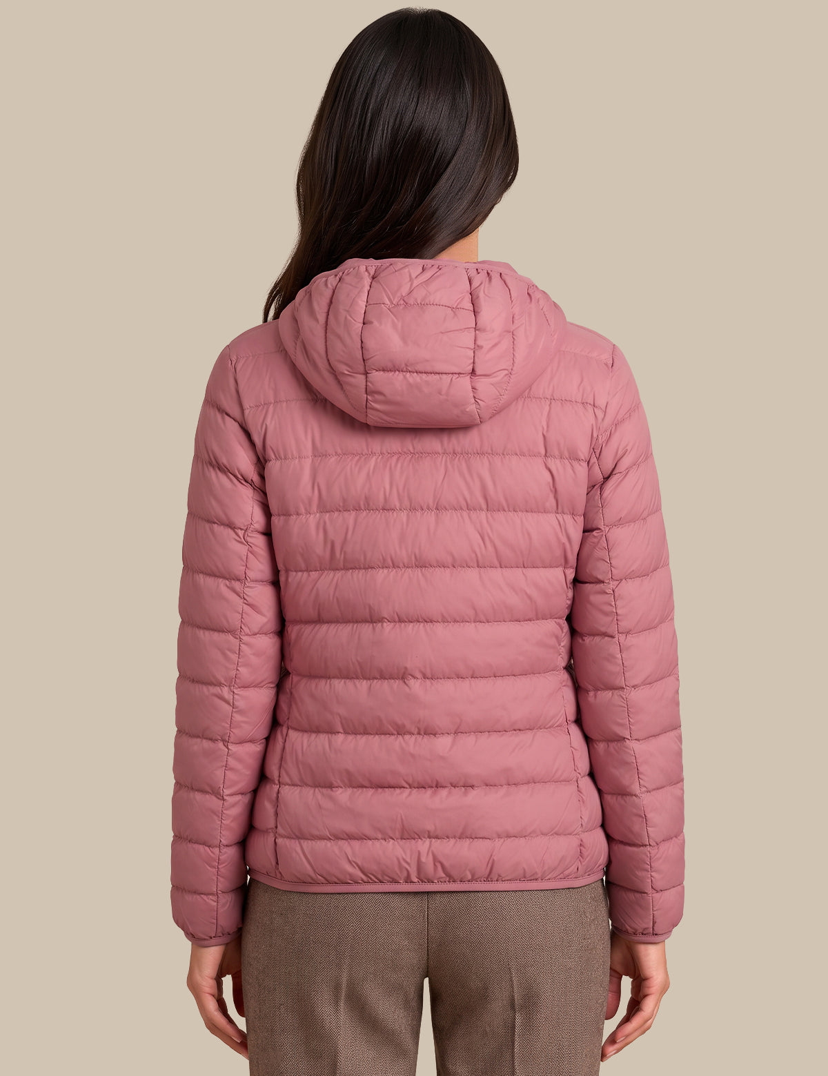 A woman viewed from the back wearing a dusty pink hooded puffer jacket and brown trousers, set against a solid beige background.