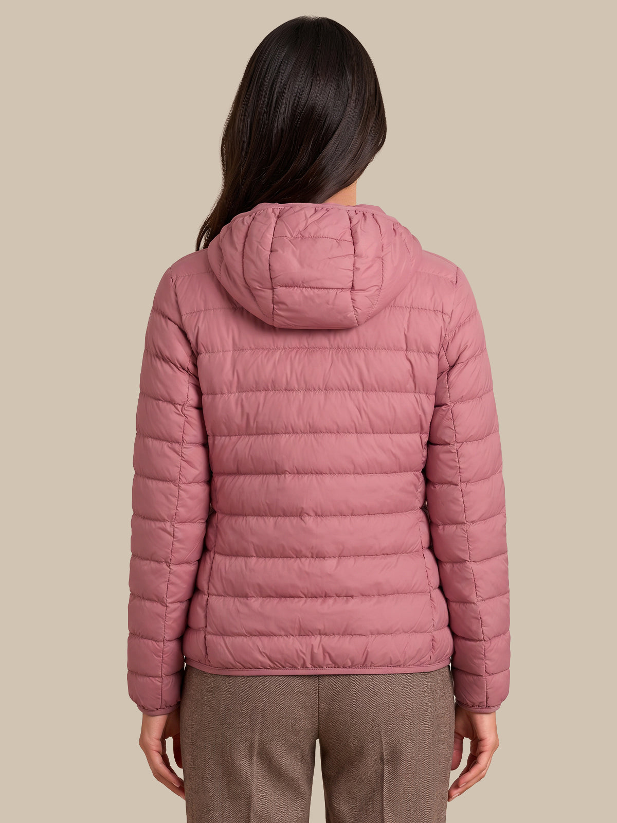 A woman viewed from the back wearing a dusty pink hooded puffer jacket and brown trousers, set against a solid beige background.