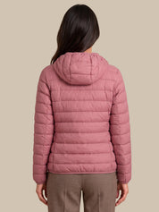 A woman viewed from the back wearing a dusty pink hooded puffer jacket and brown trousers, set against a solid beige background.