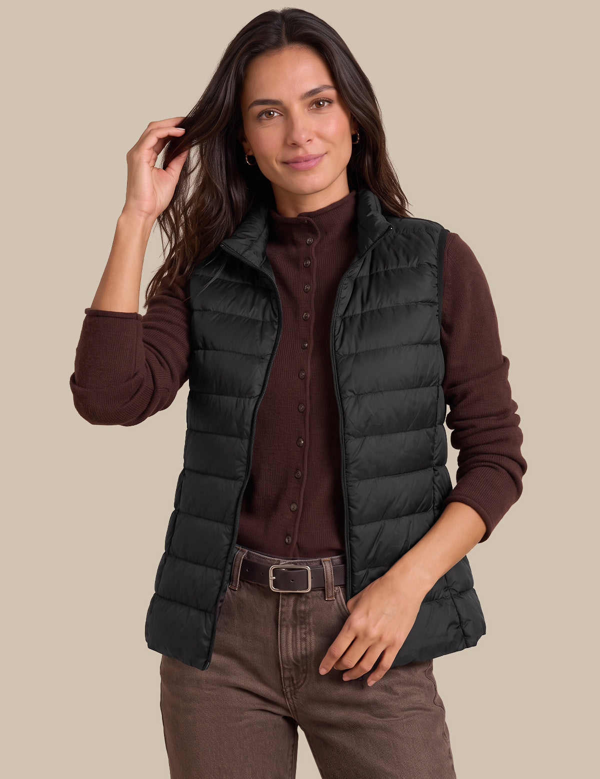 A woman modeling a black sleeveless puffer vest over a dark brown button-up shirt and brown pants, photographed against a solid beige studio background.
