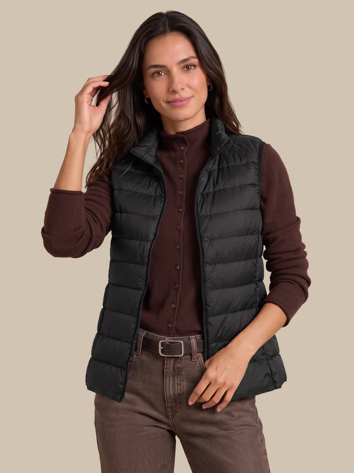 A woman modeling a black sleeveless puffer vest over a dark brown button-up shirt and brown pants, photographed against a solid beige studio background.