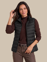 A woman modeling a black sleeveless puffer vest over a dark brown button-up shirt and brown pants, photographed against a solid beige studio background.