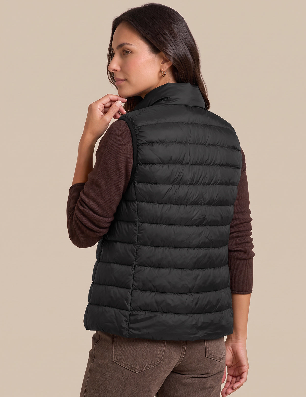 A woman shown from the back wearing a black quilted puffer vest over a dark brown long-sleeve shirt and brown pants, against a solid beige studio background.