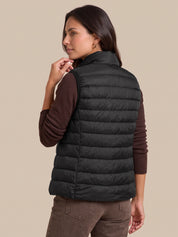 A woman shown from the back wearing a black quilted puffer vest over a dark brown long-sleeve shirt and brown pants, against a solid beige studio background.