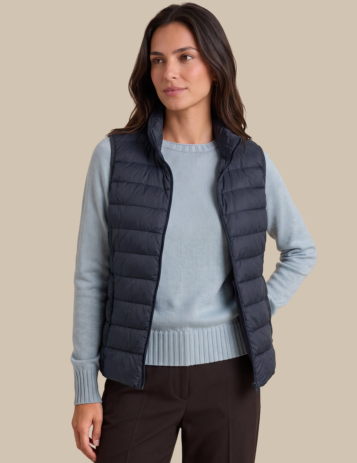 A woman wearing a dark navy quilted puffer vest over a light blue knit sweater and dark brown pants, standing against a solid beige background.