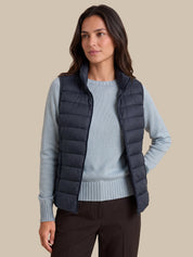 A woman wearing a dark navy quilted puffer vest over a light blue knit sweater and dark brown pants, standing against a solid beige background.