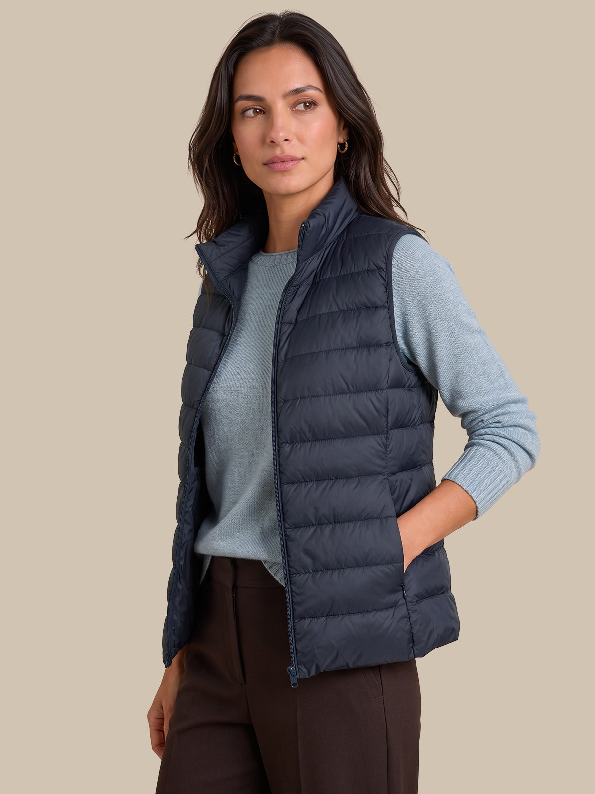 A woman modeling a navy blue puffer vest over a light blue long-sleeve top and dark brown trousers, set against a solid beige background.