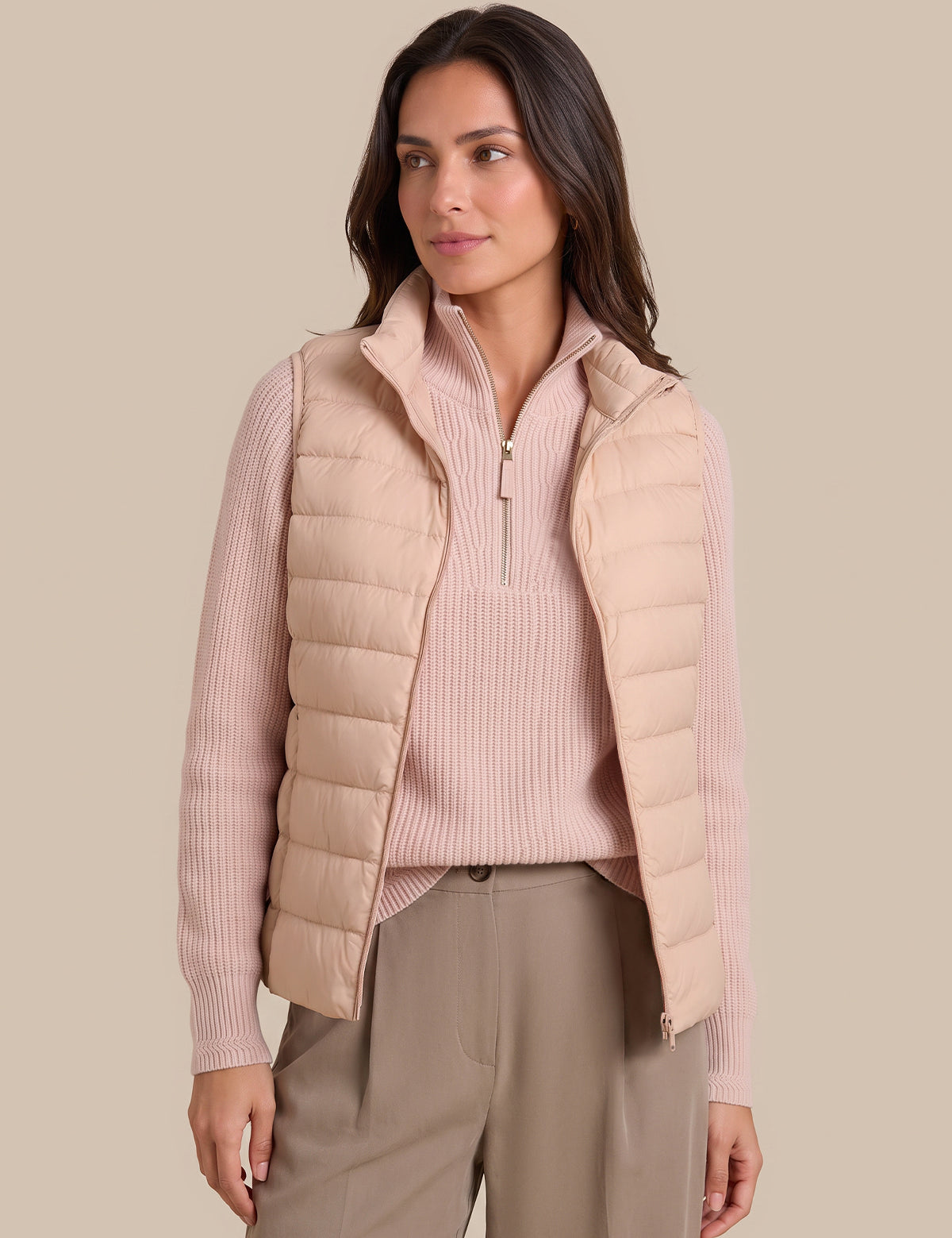 A woman modeling a light beige quilted puffer vest over a pale pink ribbed quarter-zip sweater and taupe trousers, set against a solid beige studio background.