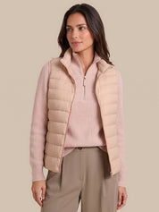 A woman modeling a light beige quilted puffer vest over a pale pink ribbed quarter-zip sweater and taupe trousers, set against a solid beige studio background.
