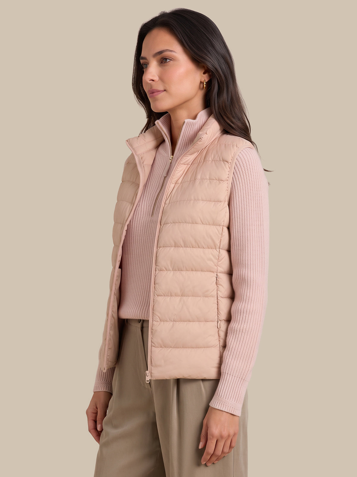 A woman wearing a light pink puffer vest over a matching pink ribbed knit top and beige trousers, photographed against a solid beige studio background.
