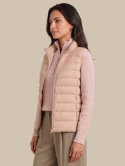 A woman wearing a light pink puffer vest over a matching pink ribbed knit top and beige trousers, photographed against a solid beige studio background.