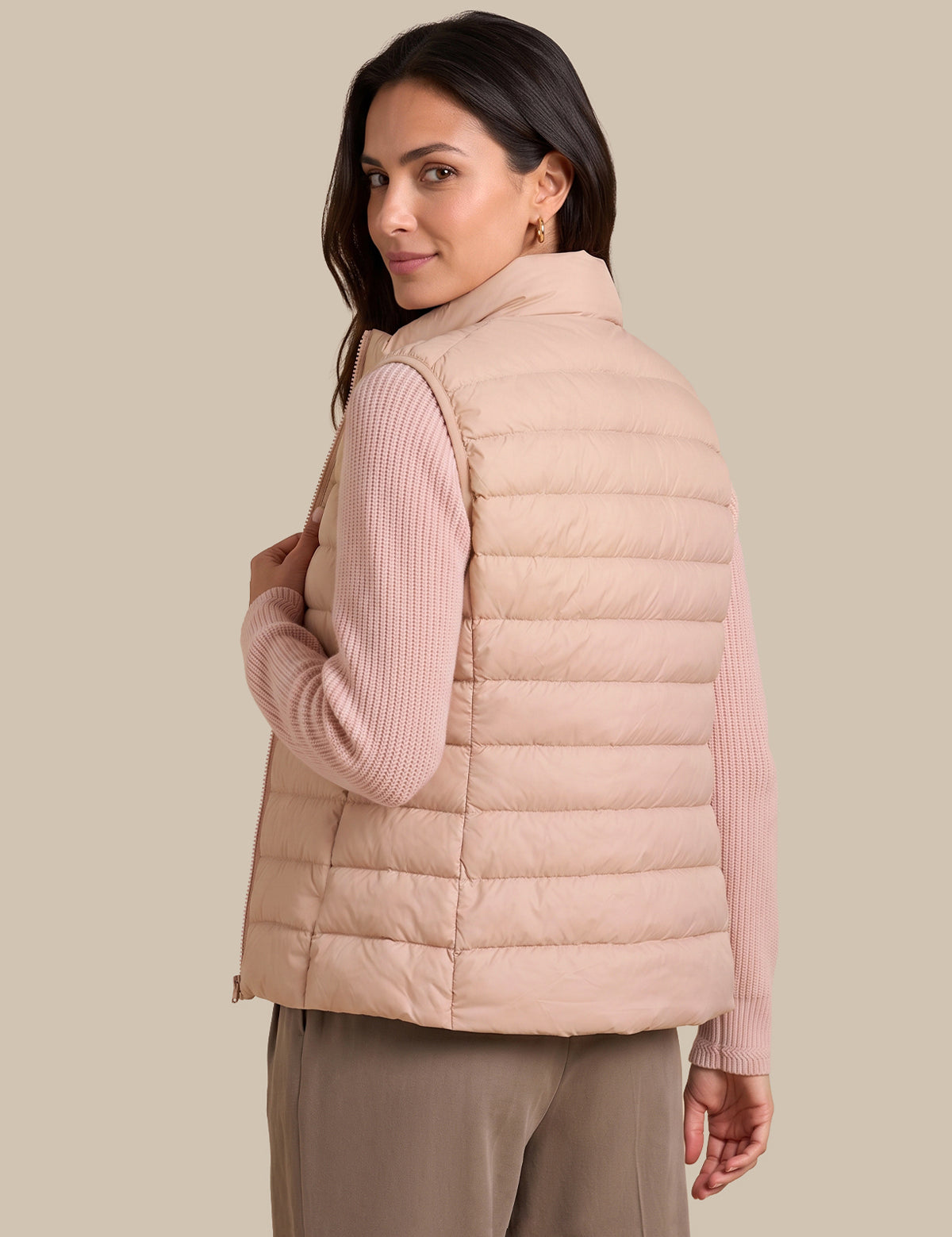 A woman wearing a light blush pink puffer vest over a ribbed long-sleeve top and taupe pants, photographed against a solid beige studio background.