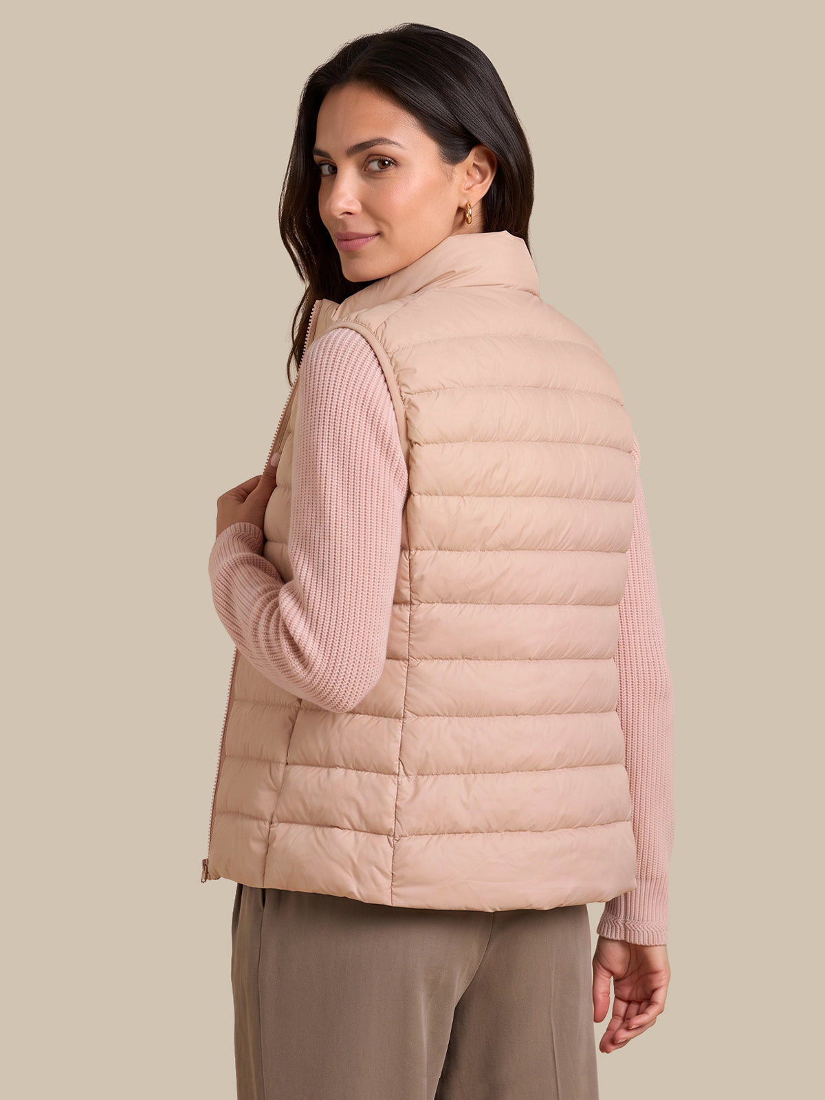 A woman wearing a light blush pink puffer vest over a ribbed long-sleeve top and taupe pants, photographed against a solid beige studio background.
