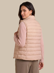 A woman wearing a light blush pink puffer vest over a ribbed long-sleeve top and taupe pants, photographed against a solid beige studio background.