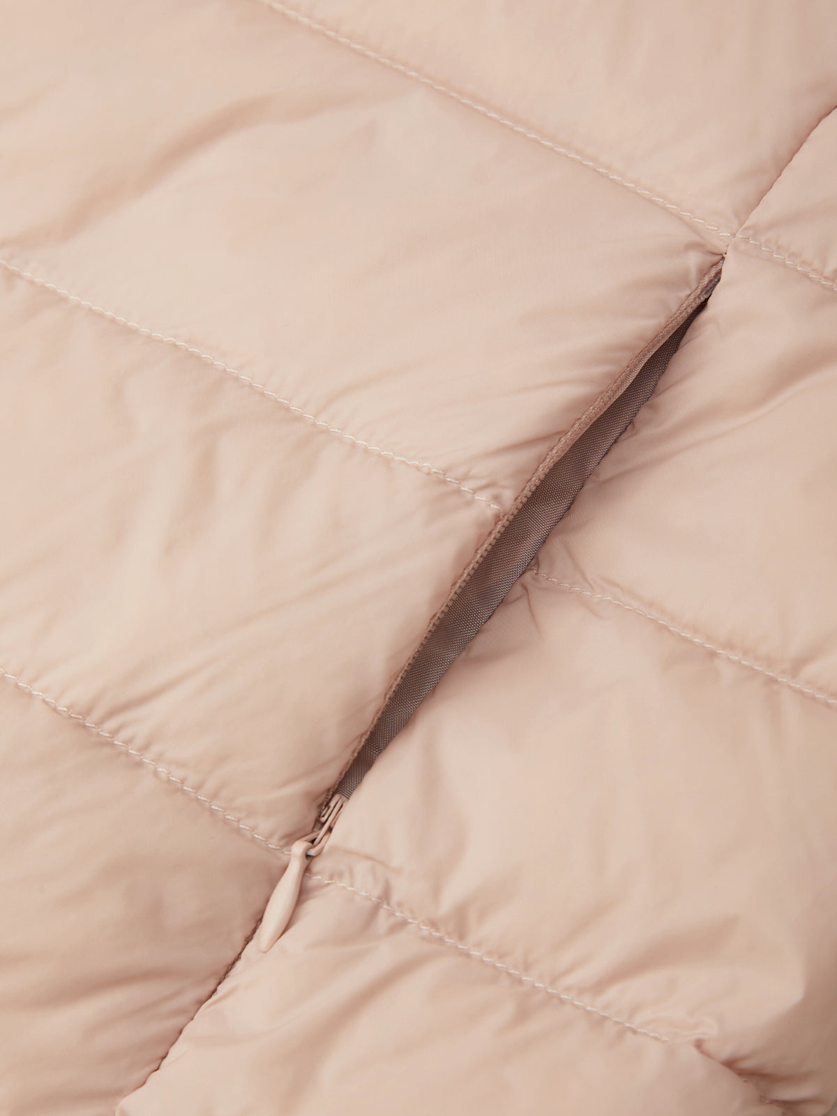 Close-up of a quilted puffer jacket in a light blush pink color, showing the texture of the fabric and a silver zipper detail.
