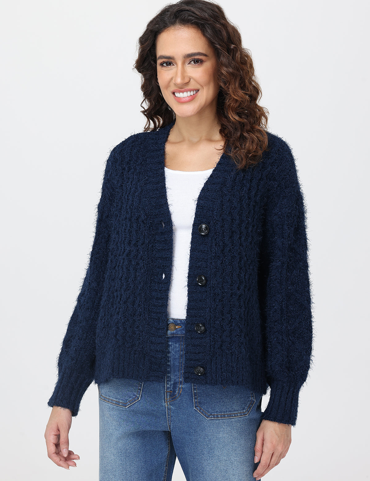 Woman wearing a navy blue cardigan over a white shirt and blue jeans on a plain background