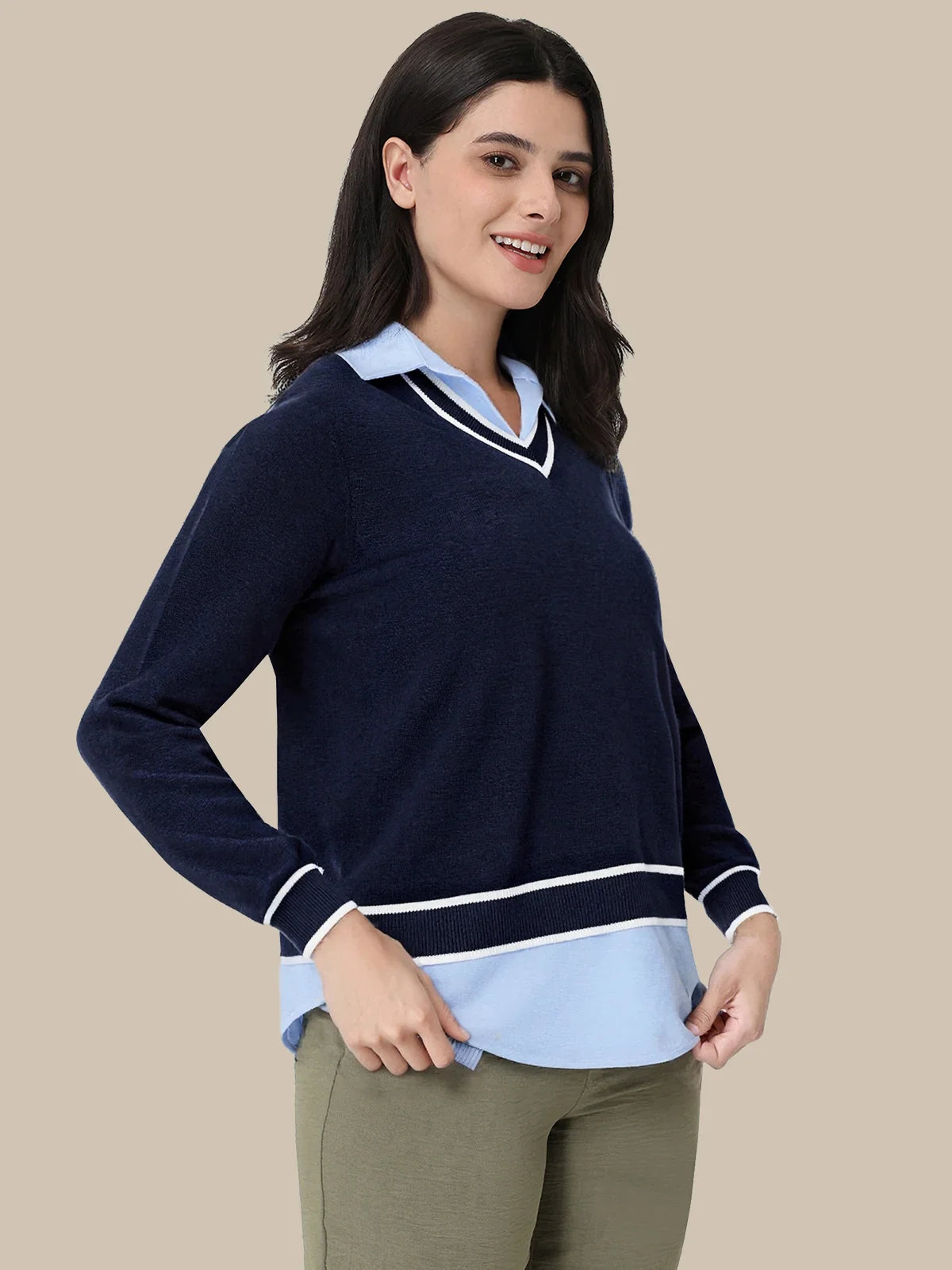 A woman modeling a navy blue long-sleeve knit sweater with a light blue and white V-neck collar and a matching light blue hem detail, paired with olive green pants, against a solid beige background.