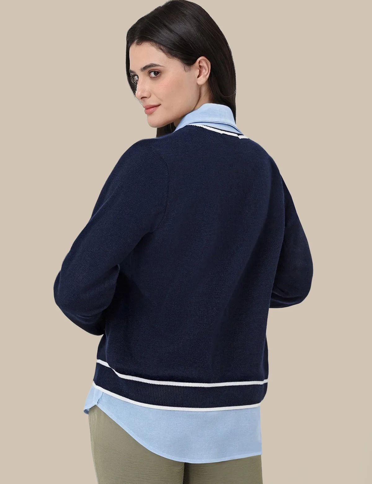 A woman shown from the back wearing a navy blue knit cardigan with white stripes, layered over a light blue collared shirt and olive green pants, against a solid beige background.