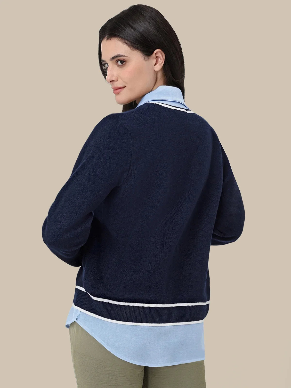 A woman shown from the back wearing a navy blue knit cardigan with white stripes, layered over a light blue collared shirt and olive green pants, against a solid beige background.