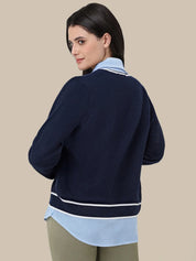 A woman shown from the back wearing a navy blue knit cardigan with white stripes, layered over a light blue collared shirt and olive green pants, against a solid beige background.