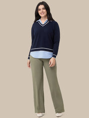 A woman modeling a navy blue long-sleeve sweater with white and light blue trim over a light blue collared shirt, paired with olive green wide-leg pants, against a solid beige background.
