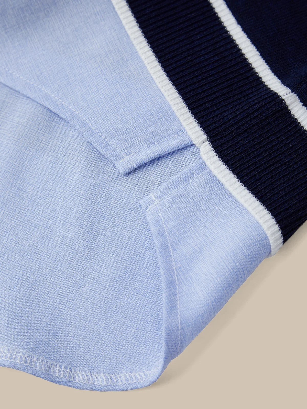 Close-up detail of a knit garment showing a light blue body, a dark navy ribbed sleeve or cuff, and a thin white stripe separating them, set against a plain beige background.