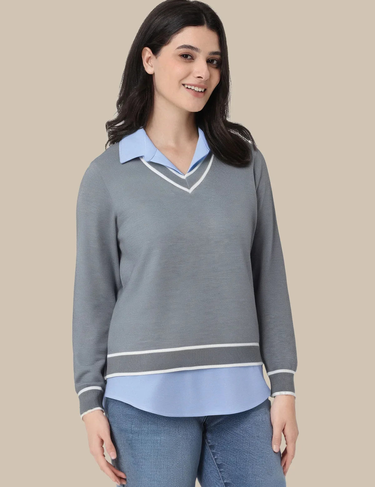A woman modeling a gray long-sleeve sweater with a light blue collar and hem, accented with white stripes, paired with blue jeans against a solid beige background.