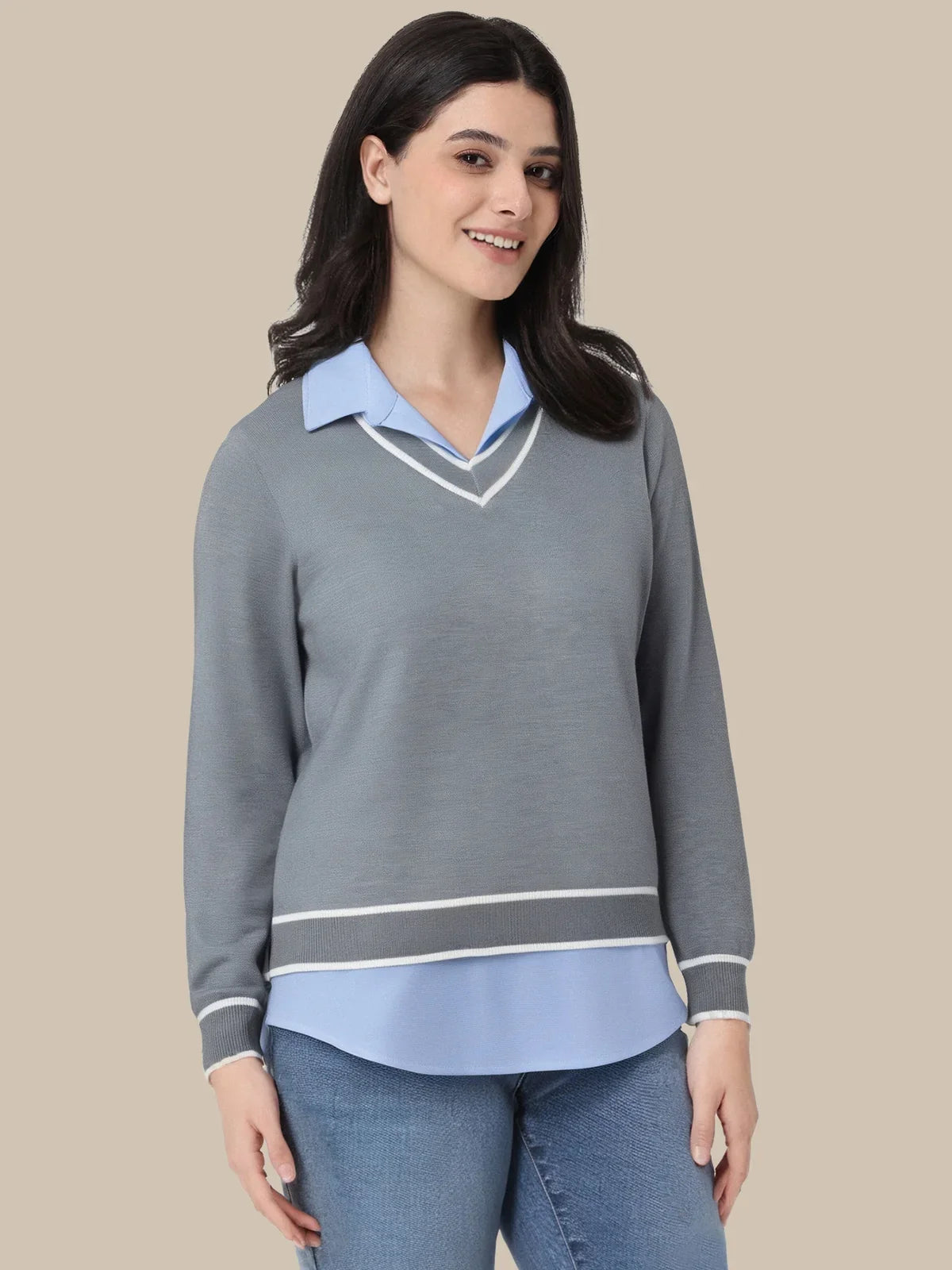 A woman modeling a gray long-sleeve sweater with a light blue collar and hem, accented with white stripes, paired with blue jeans against a solid beige background.