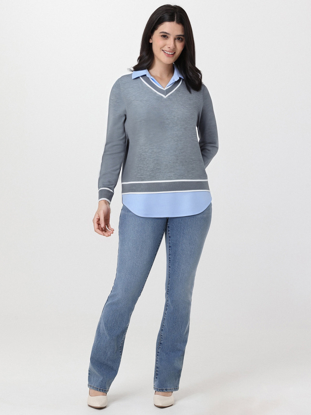 Woman wearing a gray sweater over a light blue shirt with jeans on a white background