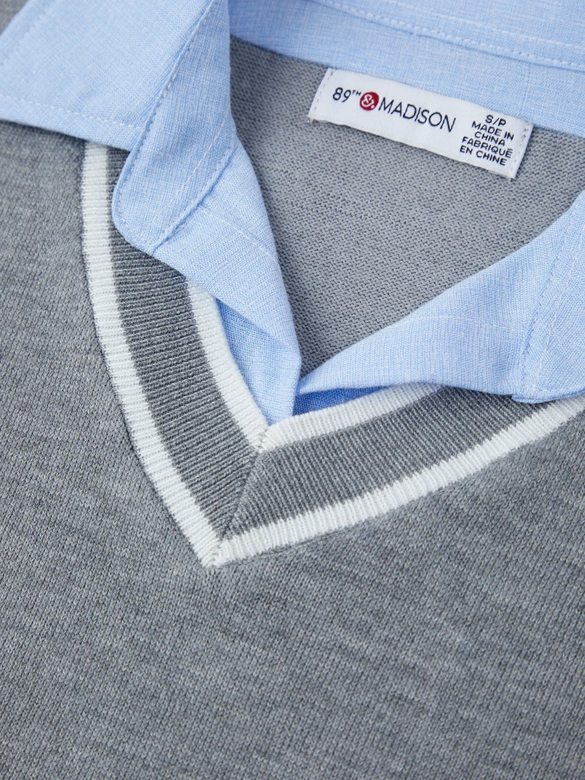 Close-up of a gray knit sweater with a light blue collared shirt layered underneath, showing a white and gray striped V-neck trim and a visible Madison 85th Street brand label.