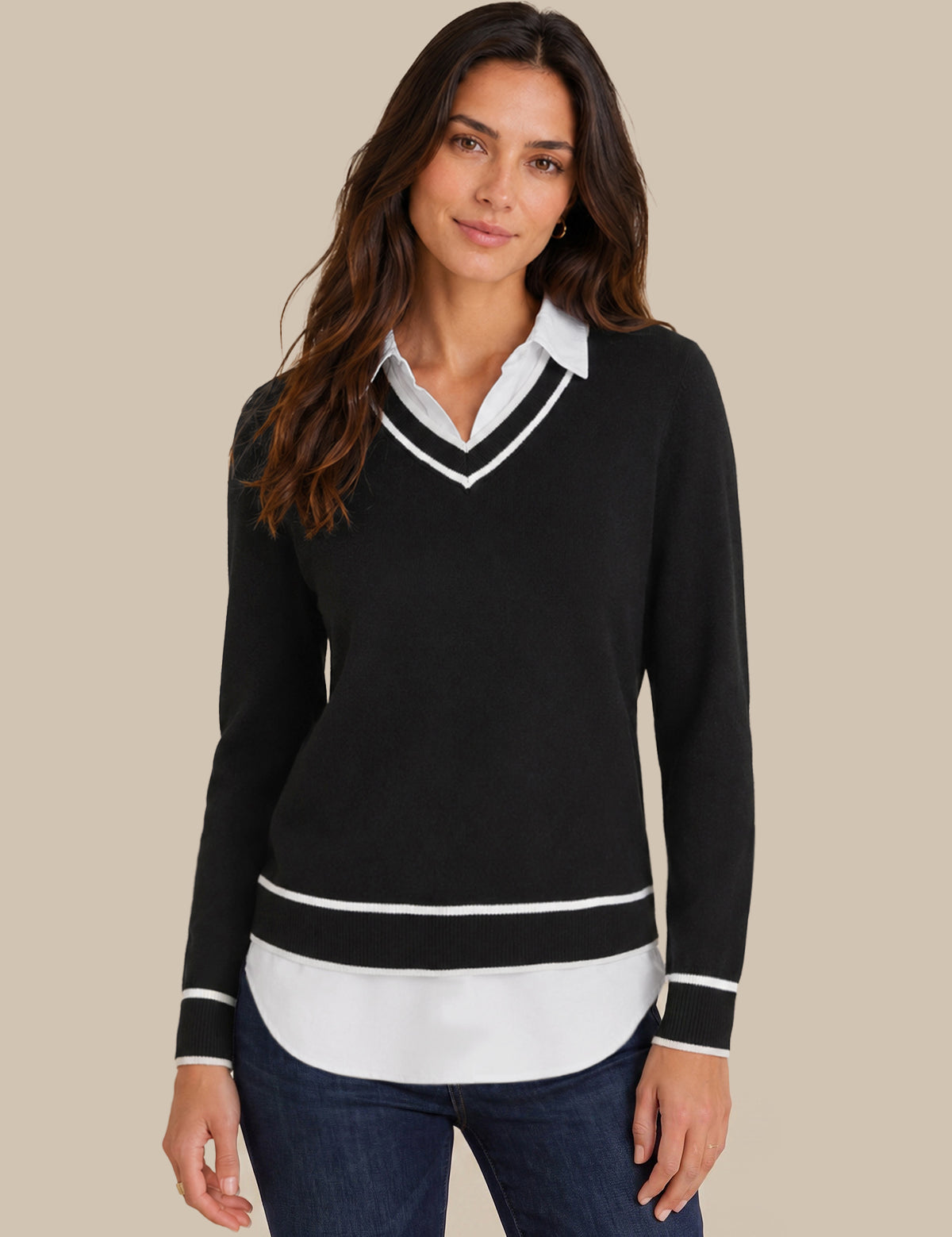 A woman modeling a black long-sleeve V-neck sweater with white trim, layered over a white collared shirt and dark blue jeans, against a solid beige background.