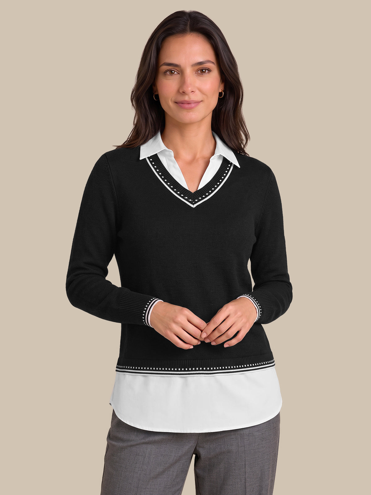A woman modeling a black long-sleeve knit sweater with a white V-neck collar trim and a white underlayer visible at the hem and collar, paired with gray trousers, against a solid beige background.