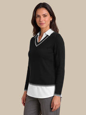 A woman modeling a black long-sleeve knit sweater with a white V-neck collar and white hem detail, styled over a white collared shirt and gray trousers against a solid beige background.