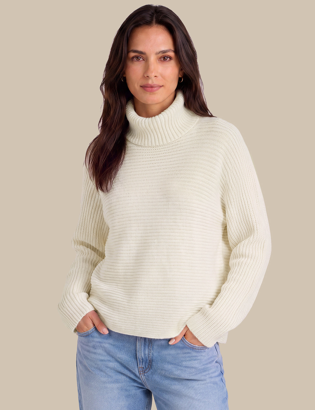 A woman wearing a cream ribbed turtleneck sweater and light blue jeans, standing against a solid beige background.