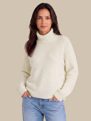 A woman wearing a cream ribbed turtleneck sweater and light blue jeans, standing against a solid beige background.