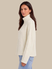 A woman wearing a cream ribbed turtleneck sweater and light blue jeans, shown from a side profile against a solid beige background.
