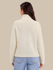 A woman shown from the back wearing a cream ribbed turtleneck sweater and light blue jeans against a solid beige background.