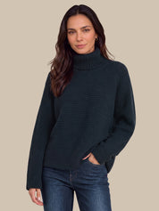 A woman wearing a dark navy ribbed turtleneck sweater and blue jeans, standing against a solid beige background.