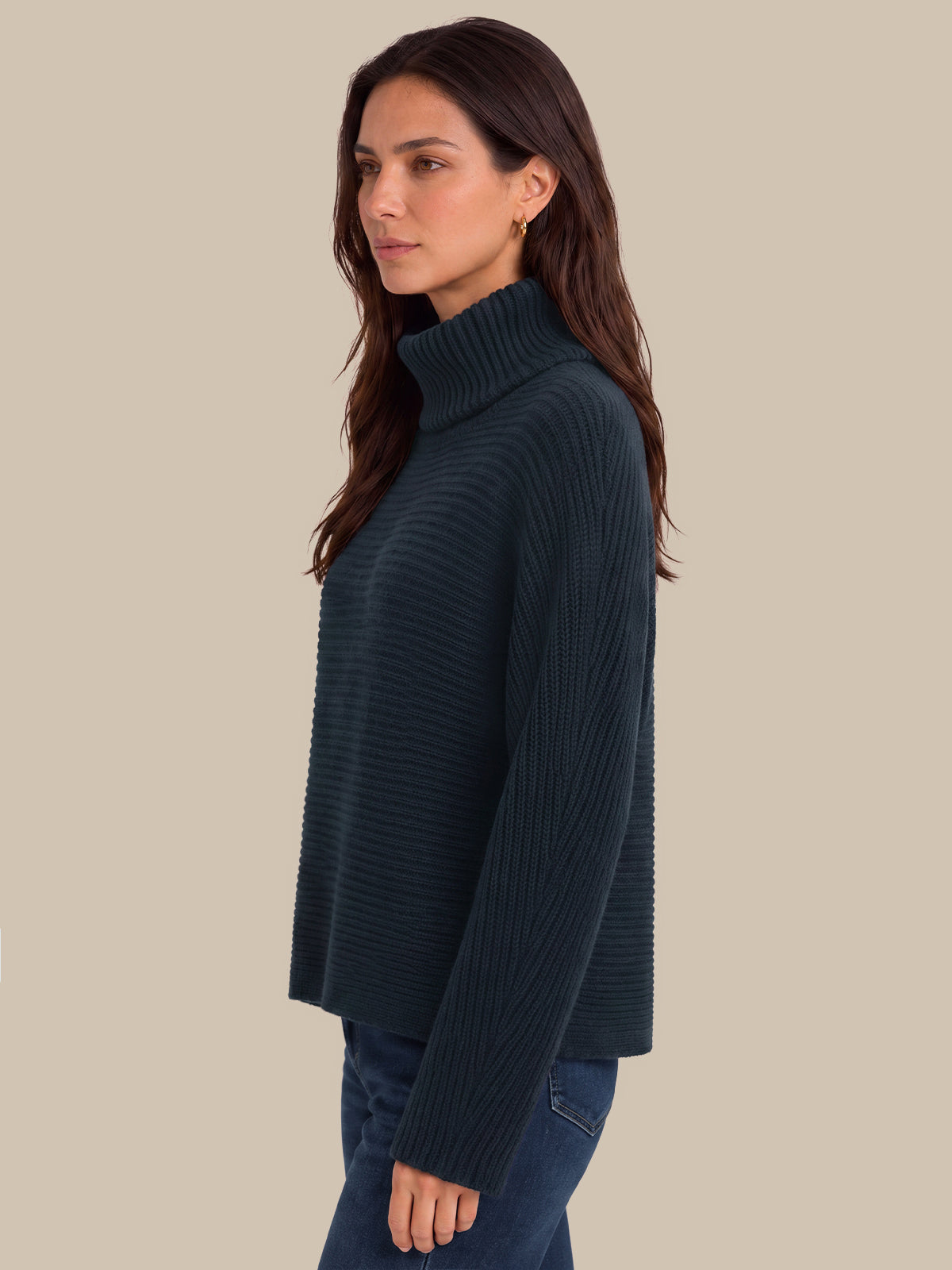 A woman shown in side profile wearing a dark navy ribbed turtleneck sweater and blue jeans against a solid beige background.