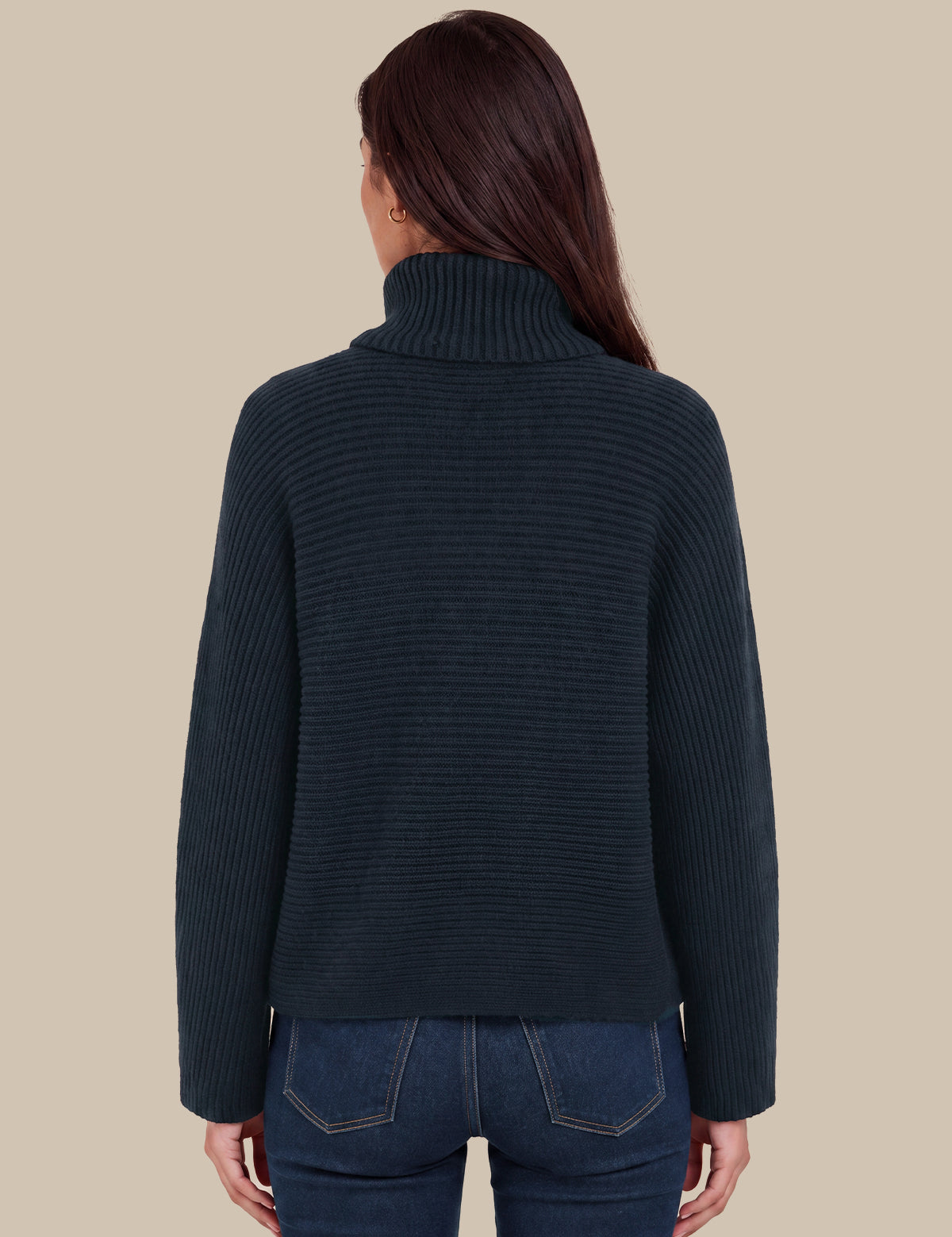 A woman viewed from the back wearing a dark navy ribbed turtleneck sweater and blue jeans against a solid beige background.