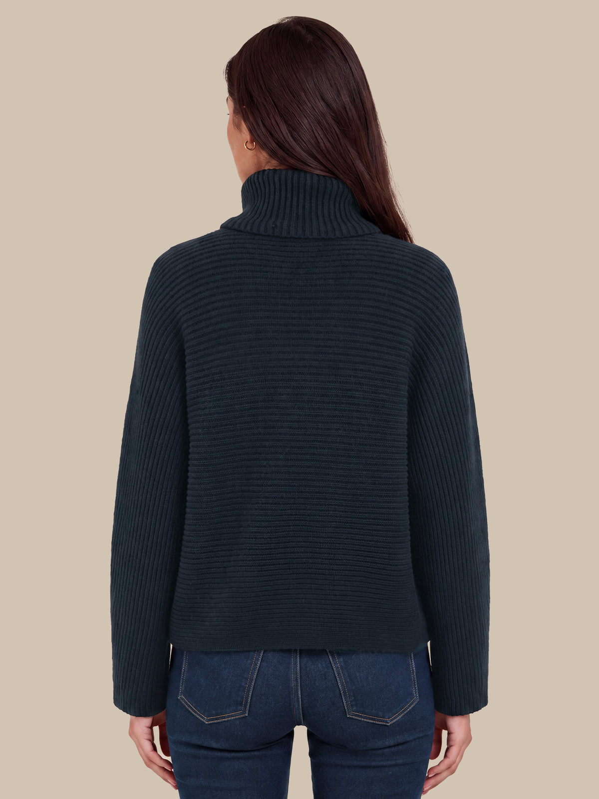 A woman viewed from the back wearing a dark navy ribbed turtleneck sweater and blue jeans against a solid beige background.