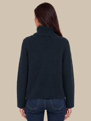 A woman viewed from the back wearing a dark navy ribbed turtleneck sweater and blue jeans against a solid beige background.