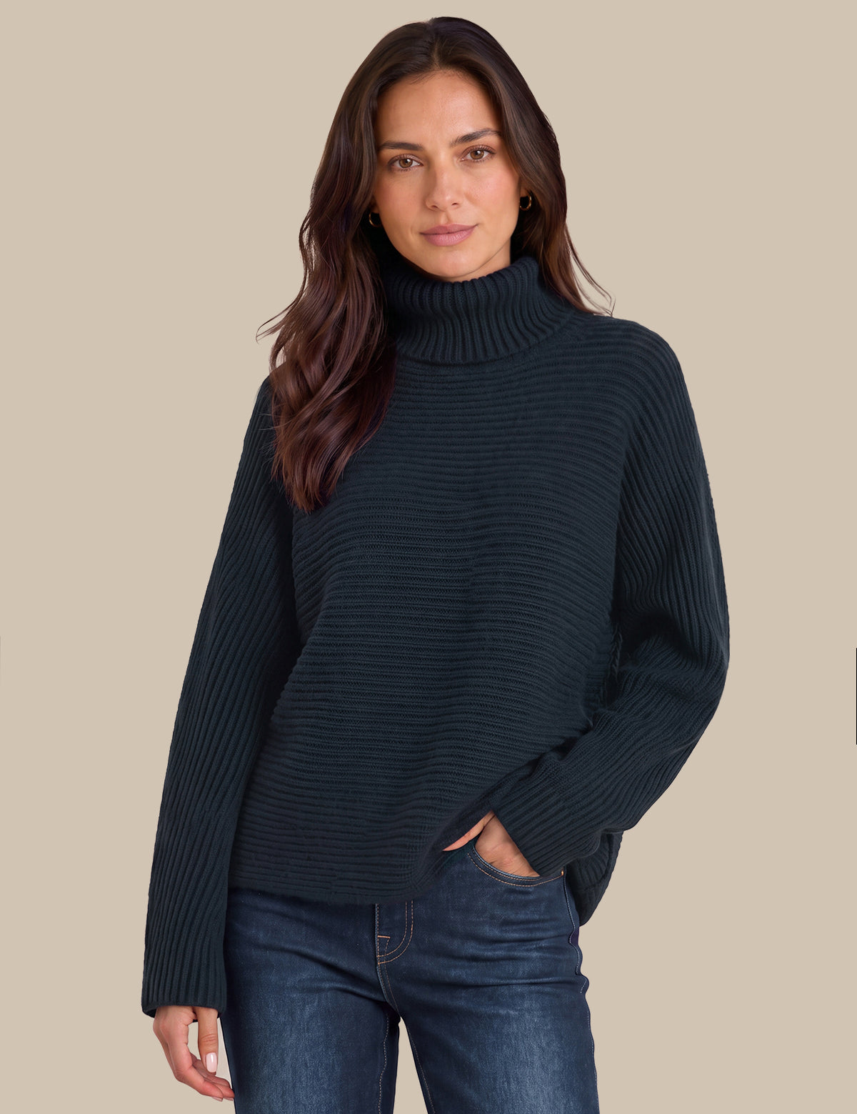 A woman wearing a dark navy ribbed turtleneck sweater and blue jeans, standing against a solid beige background.