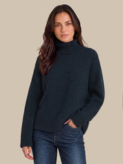 A woman wearing a dark navy ribbed turtleneck sweater and blue jeans, standing against a solid beige background.